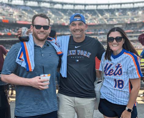 Three Media.net employees enjoying a casual outing at a baseball game, dressed in sports apparel, smiling and posing happily with the stadium crowd visible behind them.
